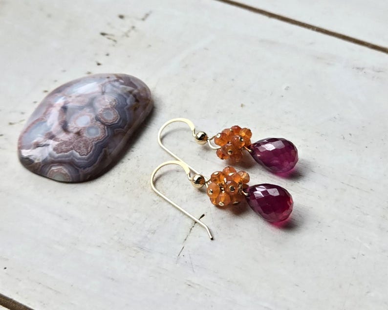 May include: Gold-tone earrings with a cluster of orange beads and a teardrop-shaped, deep pink gemstone. A polished, oval-shaped stone with swirling patterns in shades of gray, brown, and white is also present.