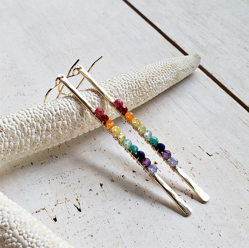 May include: A pair of gold-colored earrings with a linear design. Each earring features a row of colorful beads in a rainbow spectrum, attached to a long, rectangular gold bar. The beads include red, orange, yellow, green, blue, and purple.
