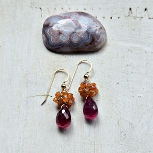 May include: Gold-tone dangle earrings featuring clusters of orange beads and teardrop-shaped, faceted, deep red gemstones. A polished, patterned stone rests in the background. Jewelry.