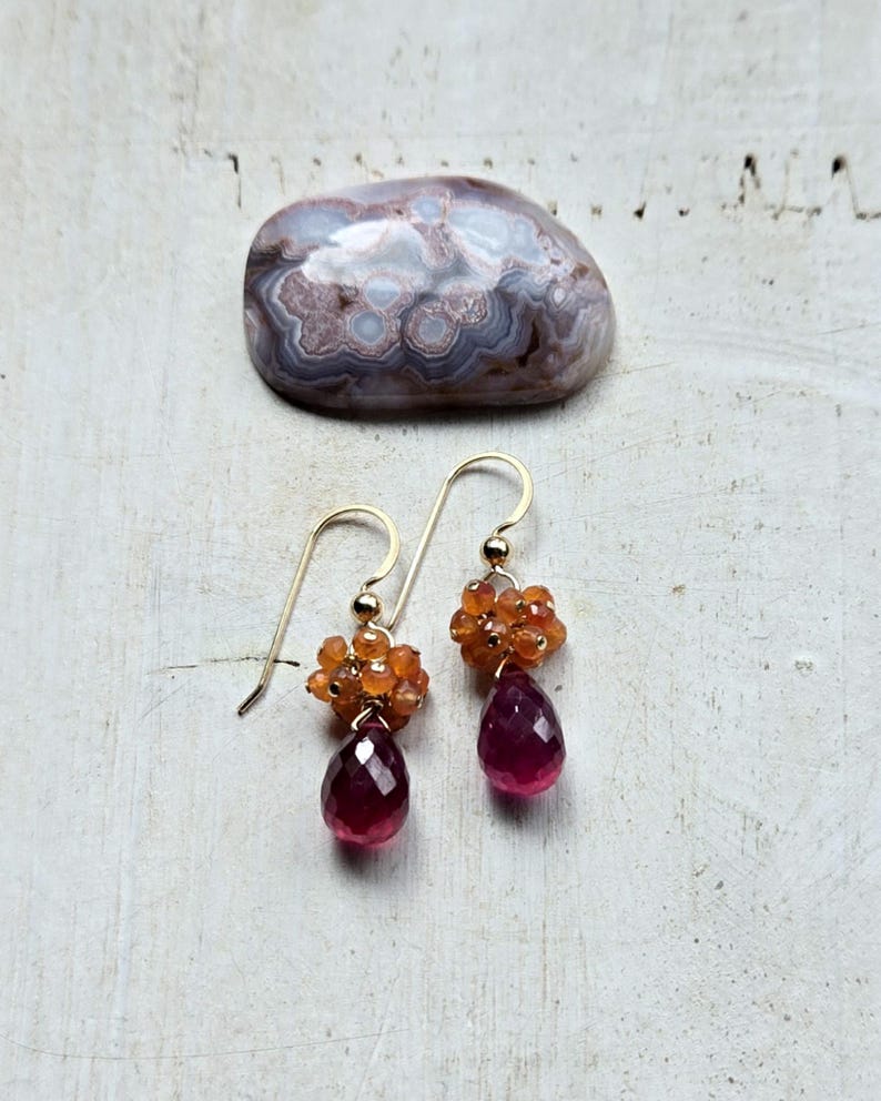 May include: Gold-tone dangle earrings featuring clusters of orange beads and teardrop-shaped, faceted, deep red gemstones. A polished, patterned stone rests in the background. Jewelry.