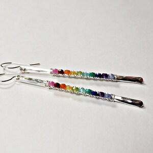May include: A pair of silver earrings with a rainbow of colorful gemstones. Each earring features a hammered silver bar with a row of small, vibrant stones in shades of red, orange, yellow, green, blue, and purple.