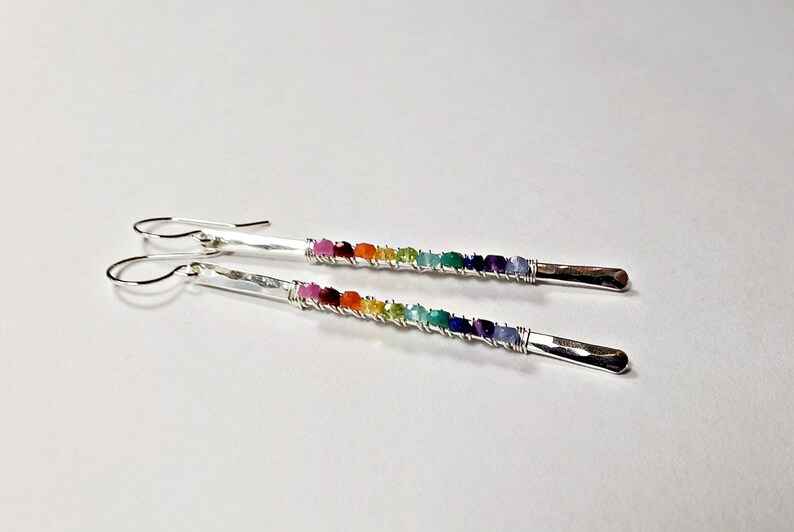 May include: A pair of silver earrings with a rainbow of colorful gemstones. Each earring features a hammered silver bar with a row of small, vibrant stones in shades of red, orange, yellow, green, blue, and purple.
