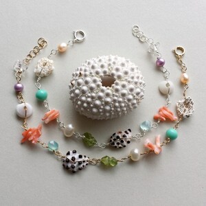 以下が含まれることがあります： ターコイズ、ピンク、ホワイトのビーズ、貝殻、コーラルのアクセントがミックスされた、繊細なネックレス2本。ネックレスは重ねて着用でき、シルバーの留め具が付いています。