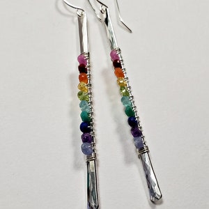 May include: Silver drop earrings with a hammered finish and a rainbow of colorful gemstone beads. The beads are strung on wire and wrapped around the silver bars. The earrings have a hook closure.