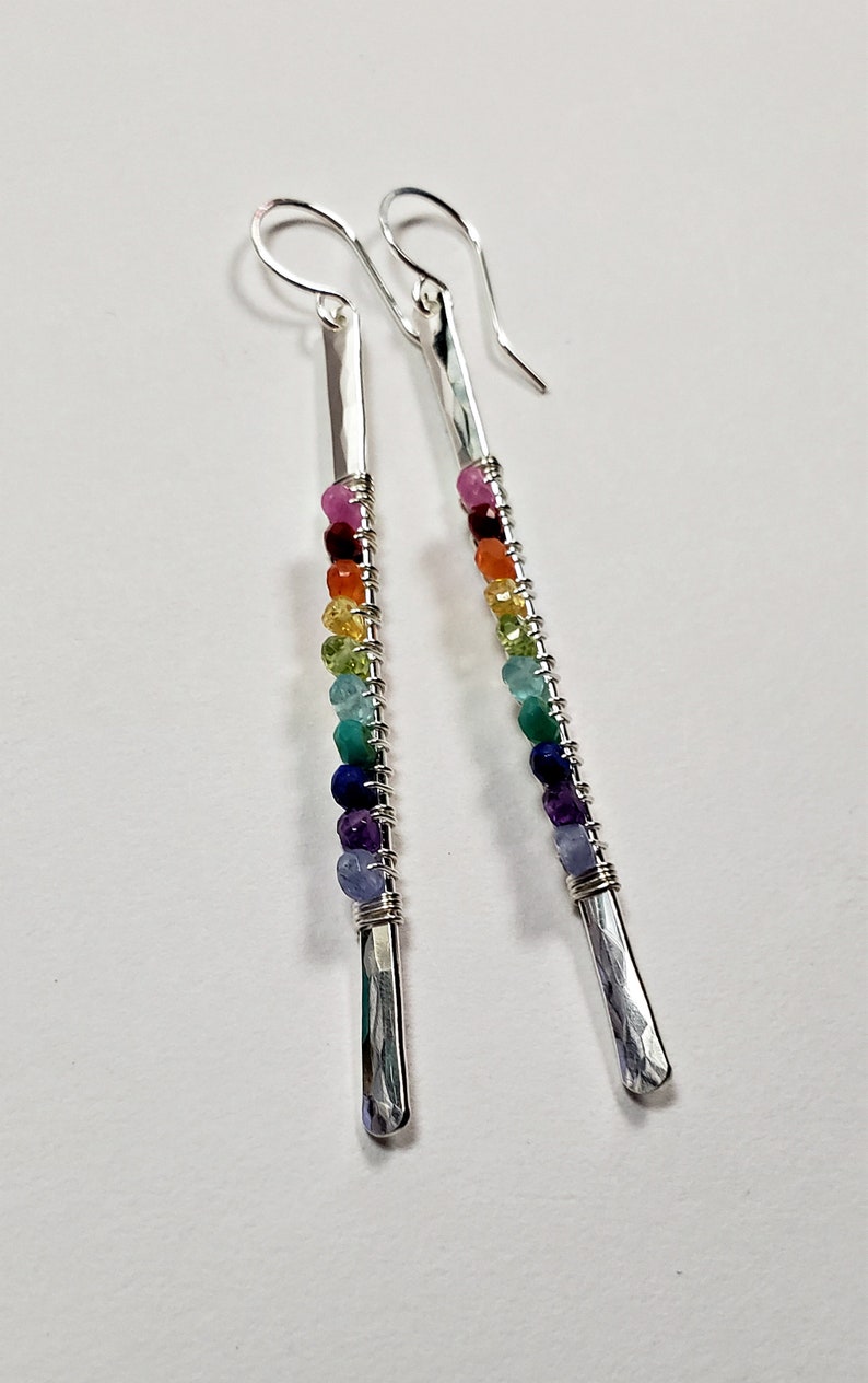 May include: Silver drop earrings with a hammered finish and a rainbow of colorful gemstone beads. The beads are strung on wire and wrapped around the silver bars. The earrings have a hook closure.
