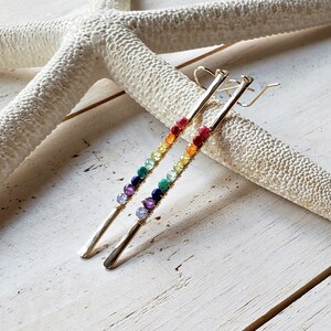 May include: A pair of gold-colored earrings with a linear design. Each earring features a series of colorful beads, arranged in a rainbow spectrum. The earrings are displayed on a white, textured surface, with a starfish in the background.
