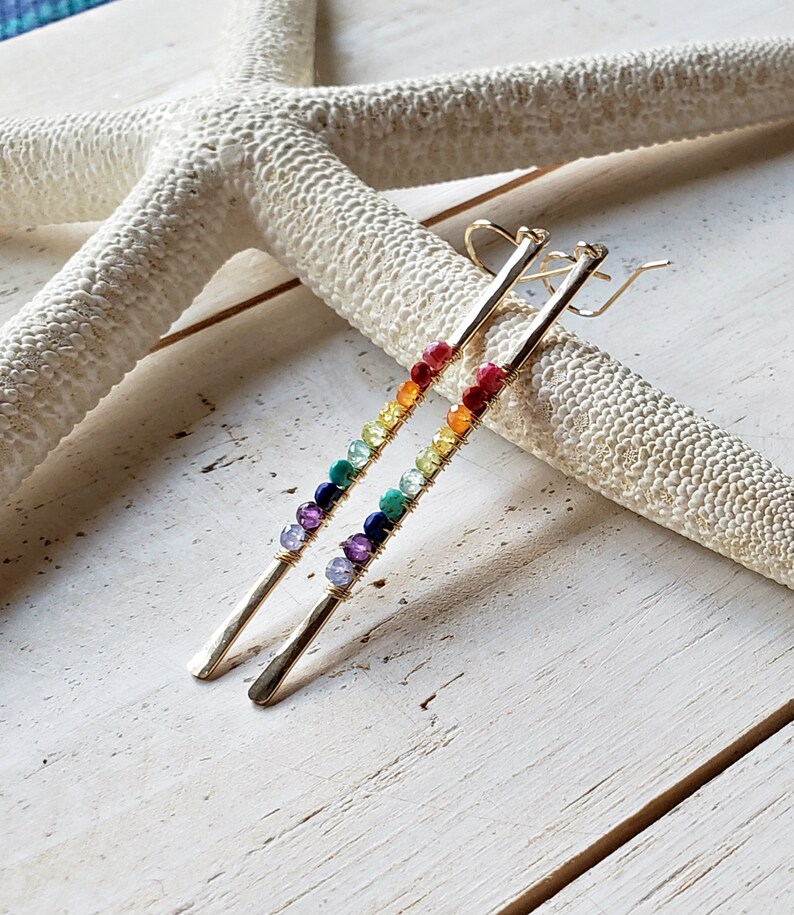 May include: A pair of gold-colored earrings with a linear design. Each earring features a series of colorful beads, arranged in a rainbow spectrum. The earrings are displayed on a white, textured surface, with a starfish in the background.