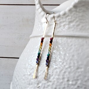 May include: A pair of gold-colored earrings with a vertical bar design. Each earring features a row of colorful beads in shades of red, orange, yellow, green, blue, and purple. The earrings are displayed against a white background.