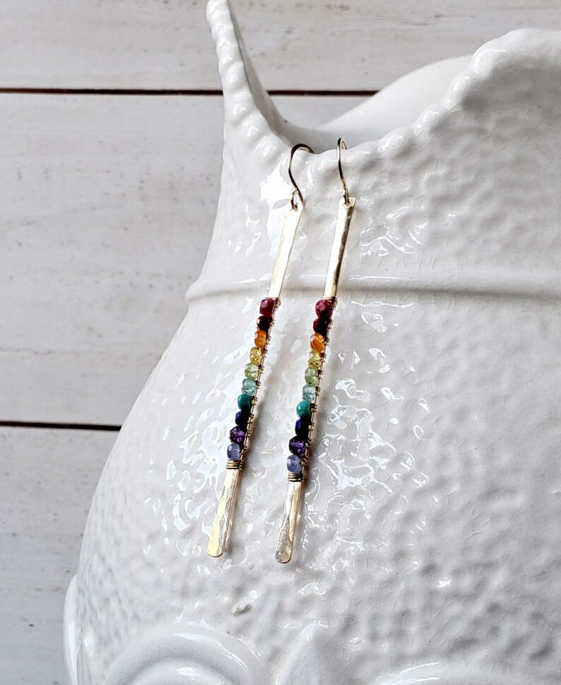 May include: A pair of gold-colored earrings with a vertical bar design. Each earring features a row of colorful beads in shades of red, orange, yellow, green, blue, and purple. The earrings are displayed against a white background.