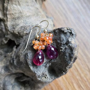 May include: Gold-tone earrings featuring clusters of orange beads and faceted, teardrop-shaped, deep pink gemstones. The earrings are displayed on a textured, gray-brown surface, showcasing their vibrant colors and intricate design.