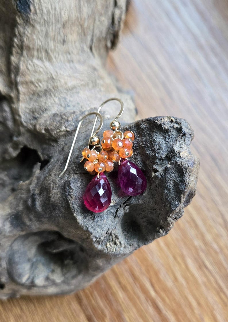 May include: Gold-tone earrings featuring clusters of orange beads and faceted, teardrop-shaped, deep pink gemstones. The earrings are displayed on a textured, gray-brown surface, showcasing their vibrant colors and intricate design.
