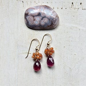 May include: Gold-tone earrings with a cluster of orange beads and a faceted, teardrop-shaped, deep red gemstone. A polished, patterned stone in shades of gray, white, and brown sits above the earrings on a white, textured surface.