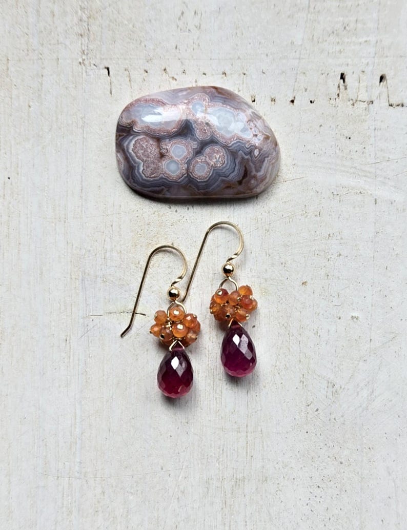 May include: Gold-tone earrings with a cluster of orange beads and a faceted, teardrop-shaped, deep red gemstone. A polished, patterned stone in shades of gray, white, and brown sits above the earrings on a white, textured surface.