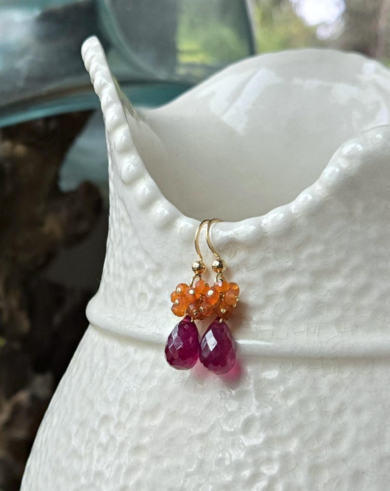 May include: Gold-tone earrings with a cluster of orange beads and two faceted, teardrop-shaped, deep red gemstones. The earrings are hanging on a white ceramic vase. The earrings are a jewelry item.