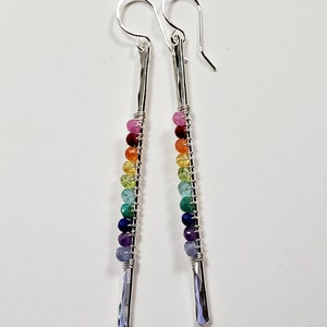 May include: A pair of dangle earrings with a rainbow of colorful beads. The earrings have a silver-colored, hammered metal bar design. The beads are strung on wire and arranged in a gradient of colors, from pink to purple.
