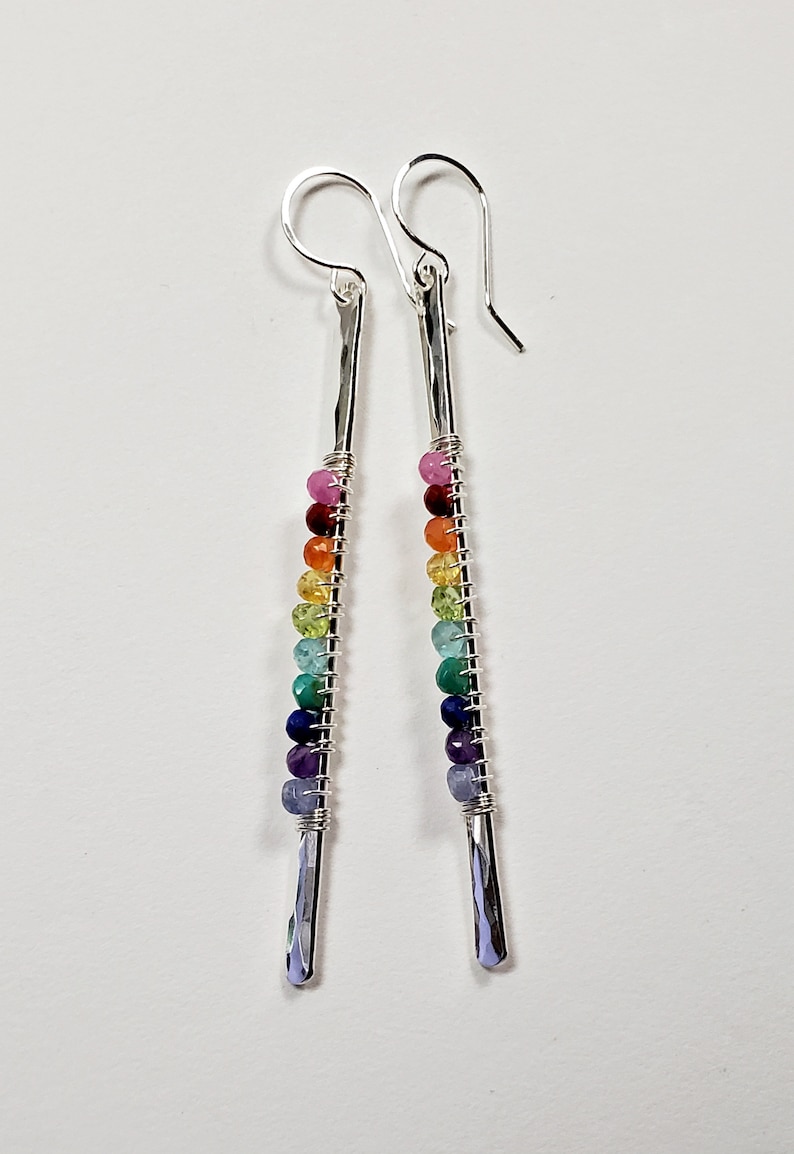 May include: A pair of dangle earrings with a rainbow of colorful beads. The earrings have a silver-colored, hammered metal bar design. The beads are strung on wire and arranged in a gradient of colors, from pink to purple.