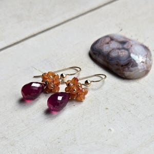 May include: A pair of gold-toned drop earrings with faceted, deep red teardrop-shaped gems. The earrings also feature clusters of small, orange-colored beads. A polished, oval-shaped stone with a mottled pattern rests nearby.