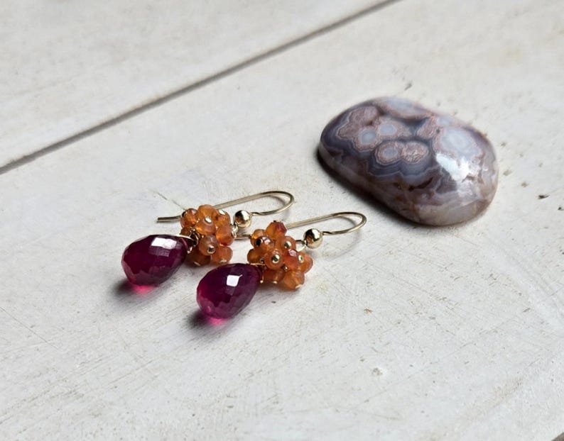 May include: A pair of gold-toned drop earrings with faceted, deep red teardrop-shaped gems. The earrings also feature clusters of small, orange-colored beads. A polished, oval-shaped stone with a mottled pattern rests nearby.