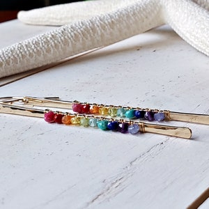 May include: Gold-tone earrings with a linear design. Each earring features a row of colorful gemstones, transitioning from red to purple. The earrings are set against a white wooden surface, with a starfish in the background.