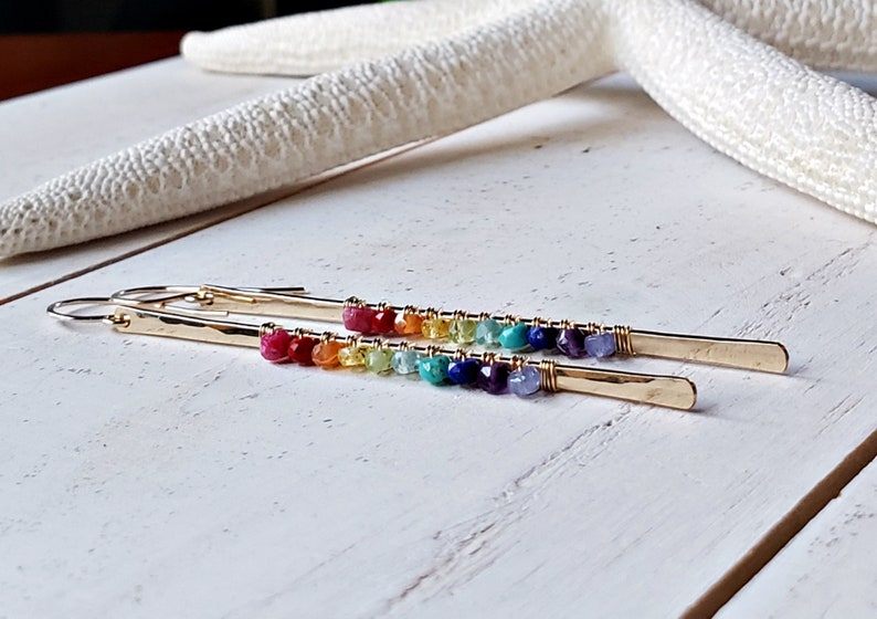May include: Gold-tone earrings with a linear design. Each earring features a row of colorful gemstones, transitioning from red to purple. The earrings are set against a white wooden surface, with a starfish in the background.