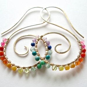 Puede incluir: Aretes de aro con piedras preciosas de arcoíris y diseño de alambre dorado. Los aretes están hechos con una variedad de piedras preciosas de colores, incluyendo rojo, naranja, amarillo, verde, azul y morado.