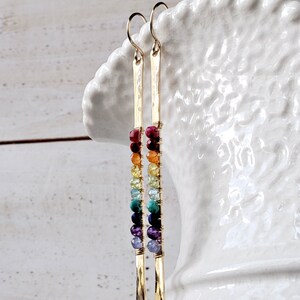 May include: Gold-tone earrings with a vertical bar design. Each earring features a series of colorful beads in a rainbow pattern, including red, orange, yellow, green, blue, and purple. The earrings are suspended from a simple hook.