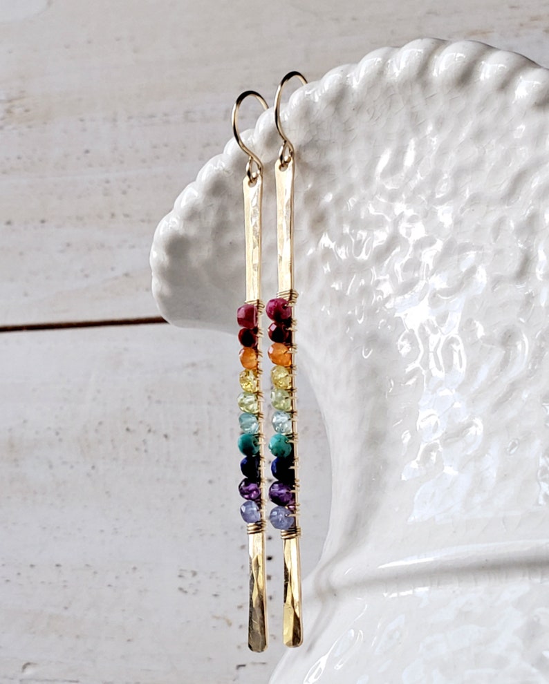 May include: Gold-tone earrings with a vertical bar design. Each earring features a series of colorful beads in a rainbow pattern, including red, orange, yellow, green, blue, and purple. The earrings are suspended from a simple hook.