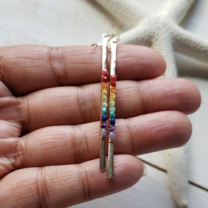 May include: A pair of gold-colored earrings with a vertical bar design. Each earring features a rainbow of small, colorful beads, including red, orange, yellow, green, blue, and purple. The earrings are displayed against a white background.