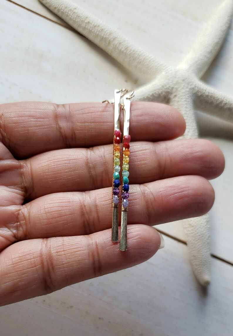 May include: A pair of gold-colored earrings with a vertical bar design. Each earring features a rainbow of small, colorful beads, including red, orange, yellow, green, blue, and purple. The earrings are displayed against a white background.