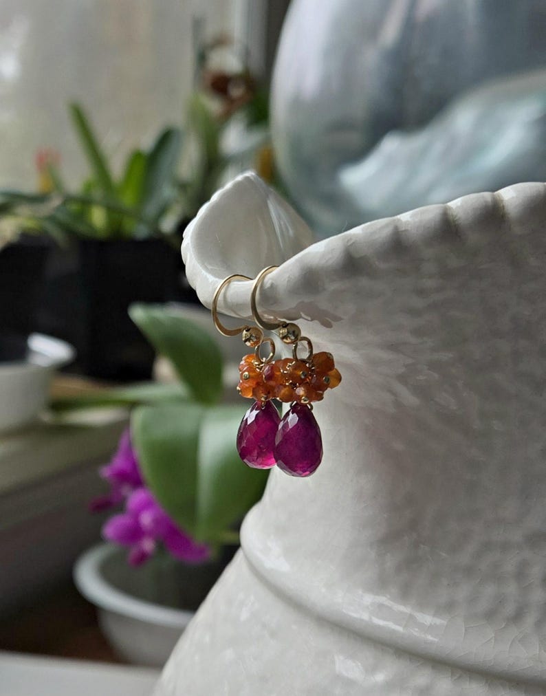 May include: Gold-tone earrings with a cluster of orange beads and two teardrop-shaped, deep pink gemstones. The earrings are displayed against a blurred background with greenery and a white ceramic vase.