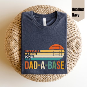 Peut inclure: T-shirt bleu marine chiné avec l'inscription "I KEEP ALL MY DAD JOKES IN A DAD-A-BASE" dans un style rétro. Le motif comprend une image stylisée d'une disquette. Le t-shirt est présenté sur un tapis tressé.