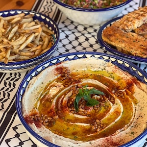 Può includere: Una ciotola bianca con un bordo blu piena di hummus condito con olio d'oliva, pinoli e paprika. L'hummus è a spirale. Un rametto di menta è posto sopra l'hummus.