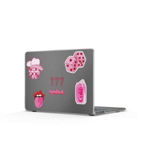 May include: A dark gray laptop lid decorated with several pink stickers, including a disco ball with a cowboy hat, two dice with hearts, a pair of lips with a tongue, a soda can labeled Coke, and the text 777 and Spoiled in pink lettering.