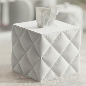 May include: A white, square tissue box with a quilted, diamond pattern. White tissues protrude from the top. The box is a solid, neutral colour, suitable for any room. The design is modern and elegant.