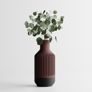 Op de afbeelding: Een moderne, geribbelde vaas in diep bordeauxrood met een zwarte basis, met een verzameling groene eucalyptus takken. De vaas heeft een cilindrische vorm met verticale groeven, waardoor een textuur ontstaat. De opstelling staat tegen een witte achtergrond.