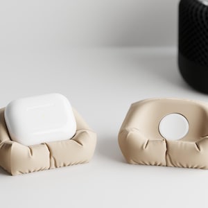 May include: Two beige leatherette charging stations, one holding a white AirPods case and the other with a white circular charging pad. The stations have a puffy, sofa-like design. A black speaker is in the background.