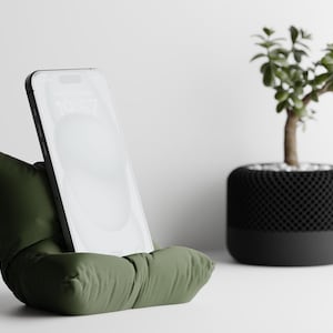 Puede incluir: Un soporte para teléfono verde oliva con un diseño similar a una almohada, que sostiene un teléfono inteligente con una pantalla brillante. Una maceta negra y gris con una pequeña planta verde en el fondo.