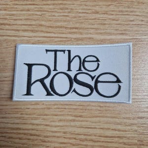 Peut inclure: Un écusson rectangulaire blanc avec le texte brodé "The Rose" en noir, avec une police stylisée. L'écusson est probablement destiné aux vêtements ou accessoires, et est posé sur un fond en bois.