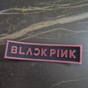 Peut inclure: Un écusson rectangulaire avec le texte "BLACKPINK" brodé en rose sur un fond noir. L'écusson est bordé de rose et est posé sur une surface sombre et texturée.