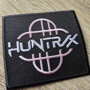 Op de afbeelding: Zwart vierkantige patch met het woord "HUNTRAX" geborduurd in wit, en een roze gestileerd ontwerp. De patch is gemaakt van een textuurstof.
