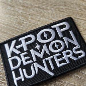 Könnte beinhalten: Schwarzer rechteckiger Aufnäher mit weiß gesticktem Text „K POP DEMON HUNTERS“. Der Aufnäher hat einen schwarzen Hintergrund und einen weißen Rand. Der Text enthält sternförmige Akzente.