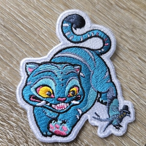 Può includere: Toppa ricamata raffigurante un gatto blu stilizzato con occhi gialli e bocca rosa. Il gatto ha un'espressione birichina. La toppa è progettata per essere applicata su abbigliamento o accessori.