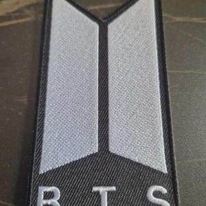 Pode incluir: Remendo retangular com borda e fundo pretos. O remendo apresenta um logotipo estilizado em branco e cinza, com as letras "BTS" bordadas em branco na parte inferior. O logotipo é um design moderno.