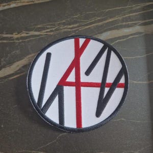 Peut inclure: Écusson rond avec un fond blanc, orné d'un motif noir et rouge. Le motif comprend un 'A' rouge et des lignes noires, le tout bordé d'un liseré noir. Idéal pour les vêtements ou accessoires.