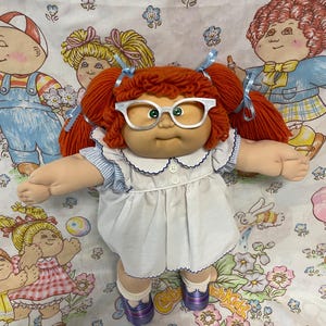 May include: A Cabbage Patch Kids doll with bright orange yarn hair styled in pigtails with light blue ribbons. The doll wears white-rimmed glasses, a white dress with a blue-trimmed collar, blue and white striped sleeves, and purple shoes.