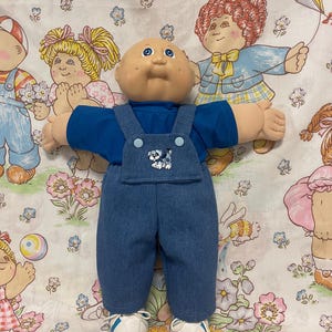 May include: A vintage Cabbage Patch Kid doll wearing a blue shirt and denim overalls with a puppy design on the bib. The doll has a bald head, blue eyes, and white sneakers. The background features a floral pattern.