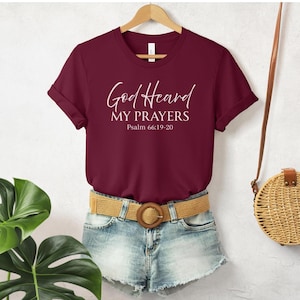 May include: A maroon t-shirt with the text "God Heard My Prayers" and Psalm 66:19-20 printed in white script. The shirt is displayed on a wooden hanger, paired with denim shorts and a tan belt. A woven crossbody bag hangs in the background.