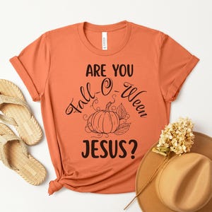 Può includere: Una maglietta arancione con la scritta "ARE YOU Fall-O-Ween JESUS?" in corsivo nero, con un'illustrazione di zucca. La maglietta è abbinata a un paio di sandali intrecciati e un cappello marrone con un fiore.