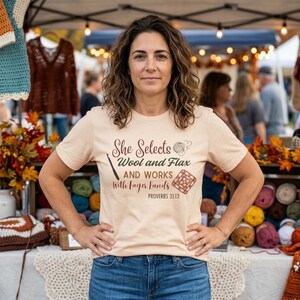 May include: A woman wearing a light tan t-shirt with the text "She Selects Wool and Flax and Works With Eager Hands" and a crochet hook graphic. The woman is standing in front of a table with yarn and other craft items.