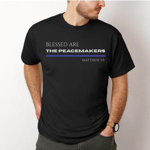 Puede incluir: Camiseta negra con el texto blanco "BLESSED ARE THE PEACEMAKERS", una línea azul y "MATEO 5:9" debajo. La camiseta es de cuello redondo.